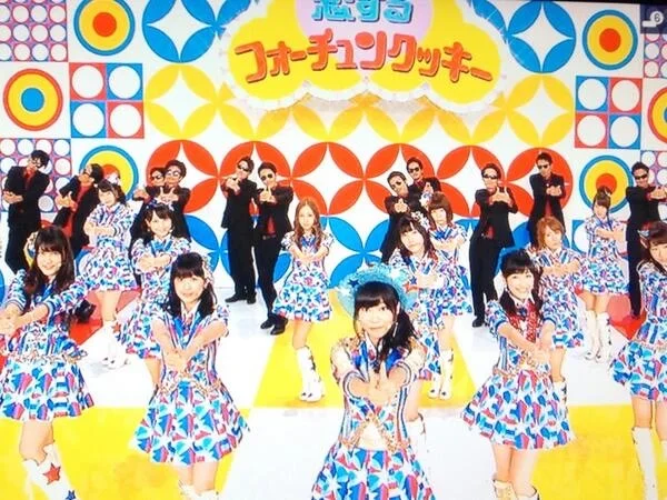 色鮮やかな衣装の人たちと、後方の黒いスーツ姿の人たちが踊るテレビ画面